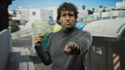 Hispanic man outdoors points at camera holding russian roubles near trash recycling containers in a...