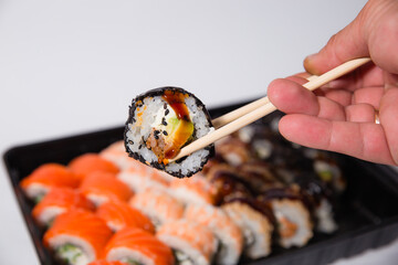 Delicious dinner, on the table various Sushi rolls with salmon, tuna, smoked eel, avocado, cream cheese laid out in a box. Sushi menu. Top view of various sushi. Holding sushi with chopsticks.