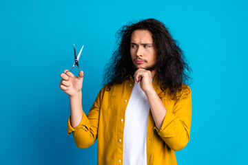 Obraz premium Young man contemplating scissors against bright blue background while wearing yellow casual shirt
