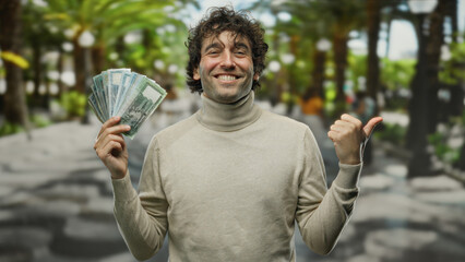 Hispanic man smiling outdoors in a city street, holding bangladesh taka banknotes and pointing...