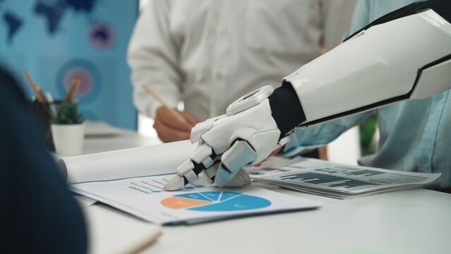 A robotic hand interacts with business reports and charts in a modern office, highlighting technology's role in teamwork and project analysis for enhanced productivity.Noogenesis
