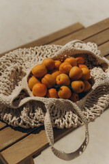 Fresh apricots in a handmade crochet mesh bag on a rustic wooden table under warm sunlight. Summer mood, eco-lifestyle, and countryside aesthetic.