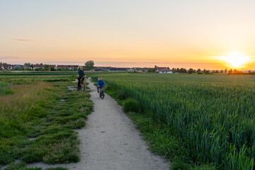 Two brothers riding bicycles on rural dirt path through green field during sunset near countryside houses. Concept of sibling bonding, childhood adventure, active lifestyle, peaceful village evening.