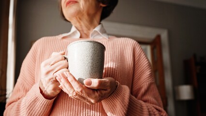 Camera view of elderly woman with stylish pink sweater holding cup of drink with both hands....