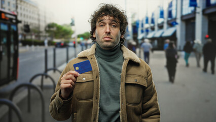 Naklejka premium Young man standing on a bustling city street holding a credit card confidently, showcasing his stylish jacket and composed demeanor in an urban outdoor setting.