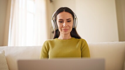 Charming Caucasian woman watching interesting video on laptop. Relaxing on sofa in cosy home. Wearing headphones while cheering for favourite streamer. Smiling sweetly. Looking at display.