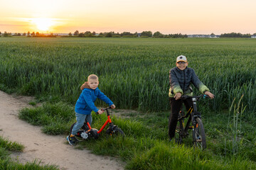 Two happy boys riding bicycles on rural path at sunset in green field. Concept of active childhood, outdoor fun and countryside adventure.