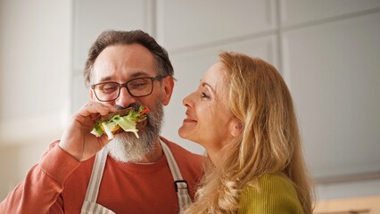Positive Caucasian woman feeding sandwich her lovely husband. Man in glasses and apron biting piece of tasty toast. Enjoying freshly prepared food. People smiling with joy in kitchen.
