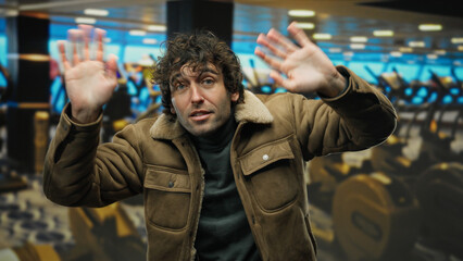 Young man with curly hair in a jacket posing indoors at a gym, showcasing a casual style with a...