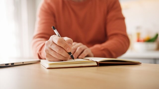Close view of man's hands writing on paper with black pen. Making notes in notebook. Bright room. Noting thoughts or plans for next month. Creativity. Being sure not to forget about task.