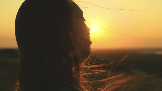 girl prays looking at sunset, long hair is flying away in glare of sunlight rays in strong wind, looking at dawn, lonely hike of brave girl, looking into sky with her eyes, believing good.