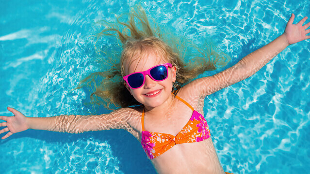 A smiling young girl wearing a bright orange bikini and pink sunglasses is floating happily in a crystal-clear blue swimming pool under the summer sun.