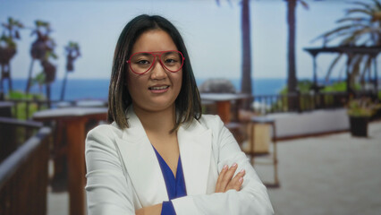 Young chinese woman in glasses and white jacket stands confidently on an outdoor terrace of a city restaurant with palm trees and ocean view in the background.