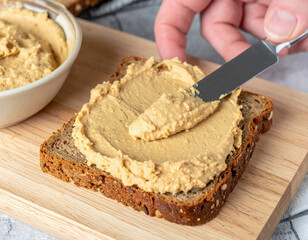 A creamy layer of hummus is being spread onto a slice of whole grain bread with a knife, highlighting a healthy and delicious snack preparation moment.