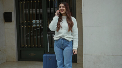 Young woman talking on phone with suitcase on street, wearing casual sweater and jeans, standing outdoors near entrance on a city sidewalk.