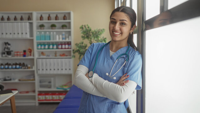 Hispanic female nurse smiling in a hospital room with arms crossed, wearing blue scrubs and stethoscope, suggesting a professional healthcare environment indoors.