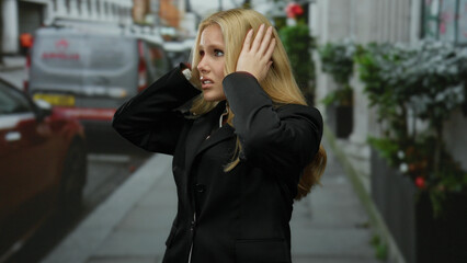 Fototapeta premium Woman outdoors covering ears in city street with vehicles and urban landscape, dressed in black suit, expressing concern, blonde hair flowing in city environment.
