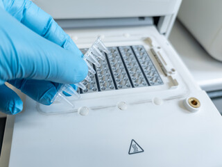 Hand with blue nitril glove holding a PCR strip with samples and PCR mix, putting a filled strip in a PCR machine. DNA analysis, biotechnology. High tech laboratory equipment. Warning sigh for heat.