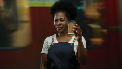 Woman in apron holding coffee cup in train station with blurred background, smiling and representing hospitality in a bustling travel environment.