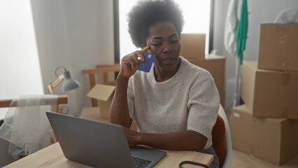 Woman using laptop in new apartment surrounded by moving boxes holding credit card looks thoughtful...