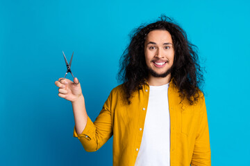 Happy young man in casual outfit holding scissors against blue background, showcasing cheerful and...