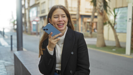 Woman recording a voice message on a smartphone while sitting on an urban street, showcasing...