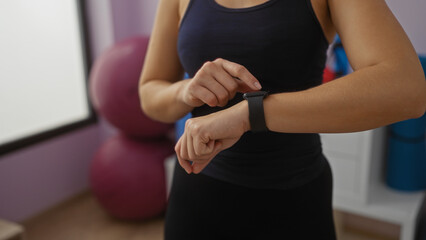 Woman adjusting smartwatch in gym interior with hands visible, highlighting active fitness lifestyle and technology integration in modern sports center environment.