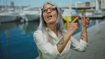Woman with grey hair points beside boats in a port, wearing glasses and a white shirt, showcasing a...