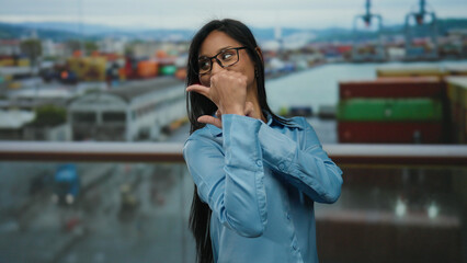 Fototapeta premium Young latin woman in glasses and blue shirt playfully poses with thumb gesture at a busy seaside port backdrop, packed with colorful containers and a blurred ocean view.