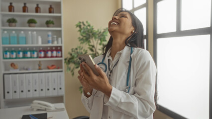 Woman wearing doctor attire smiling at phone inside clinical room showcasing a warm and professional healthcare setting with bright natural light and organized shelves.