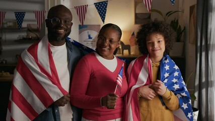 Waist-up portrait of smiling Black family consisting of married couple and pre-teen kid wrapped in American flags looking at camera in living room on independence day - Powered by Adobe