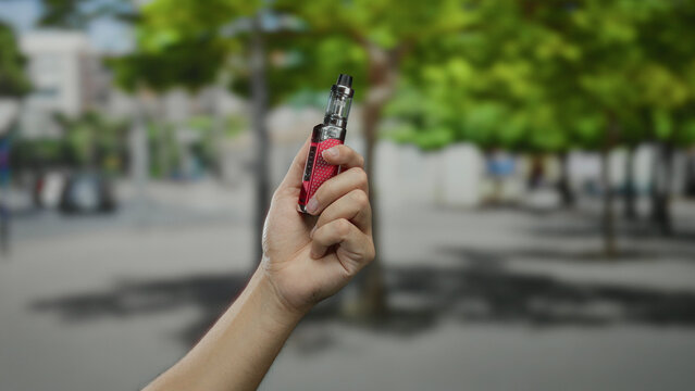 Man holds vape in hand while standing outdoors in a city street with blurred background, showcasing relaxed urban lifestyle.