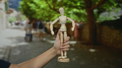 Man holding wooden mannequin on city street with blurred background of people and trees, illustrating art and creativity outdoors in urban setting.