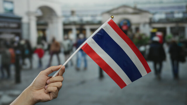 Hand holding thai flag in city street with blurred people, symbolizing thailand pride in urban setting. - Powered by Adobe
