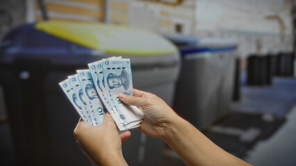 Man holding chinese yuan in front of outdoor recycling containers on a city street, suggesting...