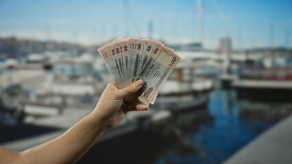 Bangladeshi man holding taka banknotes near a seaside port with boats visible in the background on...