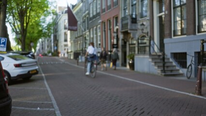 Blurred cyclist rides along a european city street surrounded by brick buildings, parked cars, and pedestrians on a sunny day, capturing urban outdoors atmosphere.