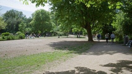 Scene in a tranquil park with blurred visitors walking on a path surrounded by lush green trees and flowers, capturing a bokeh effect with people relaxing in the distance.
