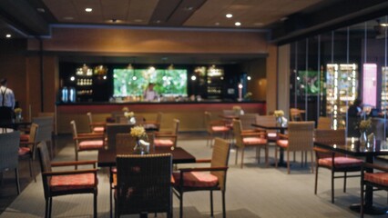 Blurred man stands at a bar counter inside an empty restaurant with tables and chairs, capturing an indoor scene with a warm atmosphere and ambient lighting.