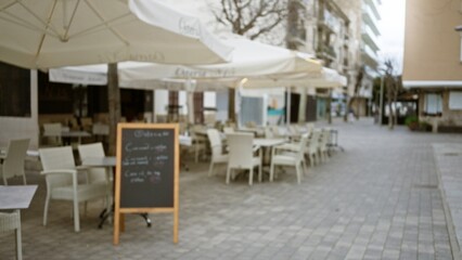 Blurred view of an outdoor cafÃ© on a cobblestone street in a mediterranean old town featuring empty chairs under large umbrellas and a blackboard menu.