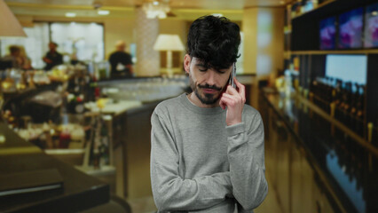 Obraz premium Young man with beard talking on smartphone in hotel bar surrounded by people and cozy lighting conveys casual ambiance and connection