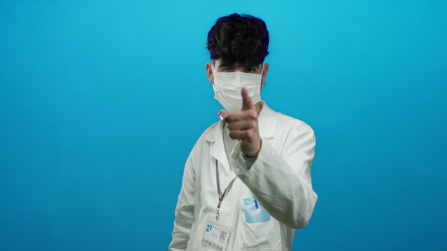 Young man in a white coat wearing a mask points at his face against a blue background, emphasizing medical awareness and responsibility.