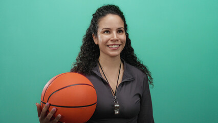 Woman holding basketball smiling against green background, showing confidence and energy, ideal for sports or coaching themes featuring hispanic and athletic vibes.