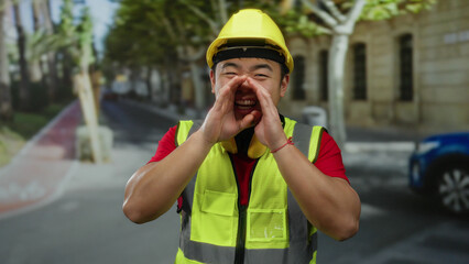 Young asian man wearing safety vest and hard hat shouting in a city street, showcasing urban...