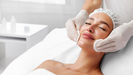 Professional cosmetologist cleansing lady's skin with cotton pads during facial beauty treatment...