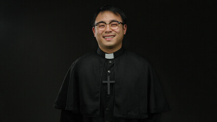 Asian priest man in glasses and black robe stands smiling against a dark background, holding hand...