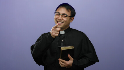 Asian man priest wearing glasses and black robe holds a bible against a purple wall, pointing...