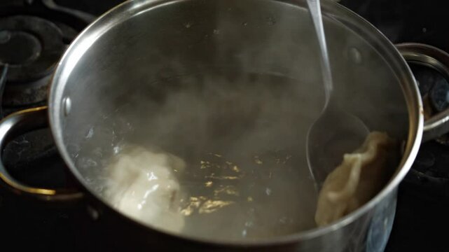 A close-up shot captures the domestic and active mood as a hand stirs dumplings or pierogi in a pot of boiling water on a gas stove, with steam rising, showcasing home cooking