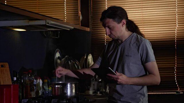 A mid-shot captures a practical and engaged mood as a man stirs a pot on a gas stove while referencing a digital tablet, illuminated by light through blinds, showing a common modern home cooking scene