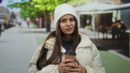 Woman dressed in a white jacket and beanie holds a coffee cup on a vibrant street, embodying a...